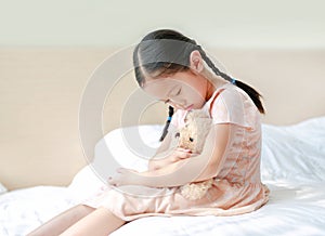 Peaceful Asian little girl embracing teddy bear while sitting on the bed at home