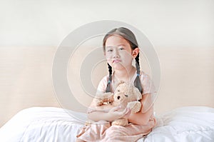 Peaceful Asian little girl embracing teddy bear while sitting on the bed at home