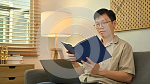 Peaceful asian man wearing glasses sitting on sofa and reading book. Leisure and people concept