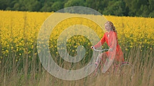 Peaceful attractive blonde lady in red dress is happily riding the bike along the blooming field covered with yellow