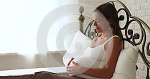 Peaceful expectant mother stroking her belly resting seated on bed