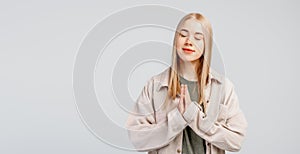 Peaceful fair-haired young woman praying with a happy smile on face, holding hands in prayer gesture and close eyes, relaxing and