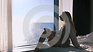 Peaceful happy young blonde woman in bathrobe enjoys her morning coffee sitting on her bed against the backdrop of