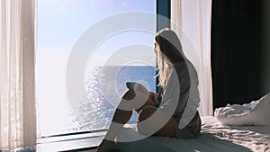 Peaceful happy young blonde woman in bathrobe enjoys her morning coffee sitting on her bed against the backdrop of