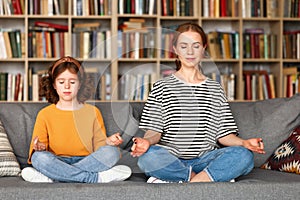 Peaceful mom and daughter meditating together while sitting in lotus pose on sofa at home