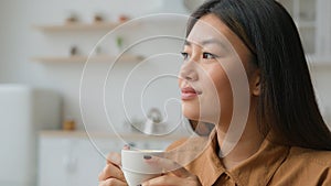 Peaceful relaxed Asian chinese japanese woman sit in kitchen hold cup of tea dreaming think feel harmony enjoy drink