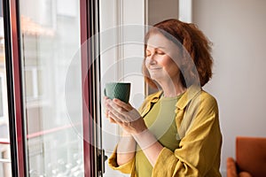 Peaceful senior woman inhaling tea aroma while standing near window at home