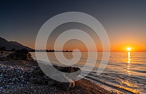 Peaceful sunset over the ocean with silhouette of mountains and pebble beach iwth rock boulders in the foreground