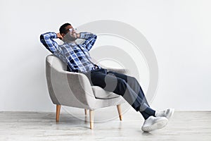 Peaceful young black man relaxing in armchair against white studio wall, free space