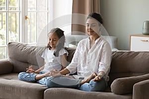 Peaceful young Indian mother teaching girl to meditate at home