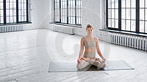 Peaceful young woman in stylish tracksuit sits on mat in full lotus yoga pose meditating