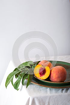 Peach in halves with bone in minimalistic style. Peaches with leaves on color green plate on table with white tablecloth. Ripe