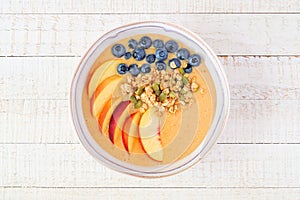 Peach smoothie bowl with blueberries and granola, top view over white wood