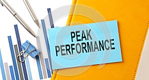 PEAK PERFORMANCE text on sticker on the yellow notebook with chart and pen