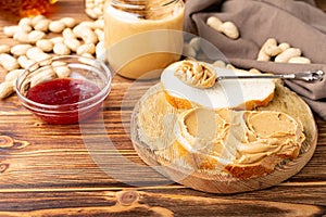 Peanut butter in spoon near creamy peanut paste in open glass jar, slice of peanut butter bread, toast, jam. Peanuts in peel