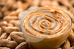 Peanut butter swirls in a glass bowl over raw peanuts background. Creamy smooth peanut butter in jar