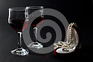 Pearl Necklace and Golden Ring in Jewelry Box with Two Wineglasses Filled with Red Wine Isolated on Black