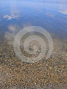 Pebbles at the bottom of a clear lake. The sky with clouds is reflected in the water