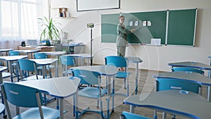 Pedagogue in empty classroom with ruler in hand near blackboard using cellphone video camera recording herself during