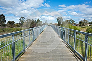 A pedestrian footbridge/boardwalk over wetlands leads to an Australian neighbourhood with some residential houses in the distance