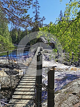 Pedestrian suspension bridge over the Ahvenkoski waterfall on the Tokhmayoki River in Karelia leading to a wooden house on a clear