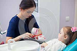Pediatric dentist educating a smiling little girl about proper tooth-brushing, demonstrating on a model. Early