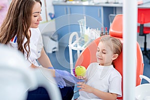 Pediatric dentist gives a little girl a big green apple. beautiful girl is smiling in dentist`s office. concept is a children`s