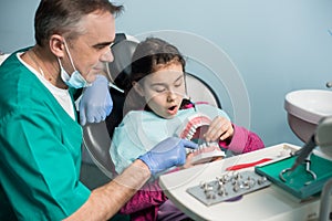 Pediatric dentist showing to girl in dentist chair dental jaw model at dental clinic