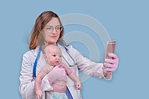 Pediatrician doctor holding phone and newborn baby, blue studio background. Happy nurse in uniform with a child and a mobile phone