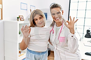 Pediatrician woman working at the clinic with little girl doing ok sign with fingers, smiling friendly gesturing excellent symbol