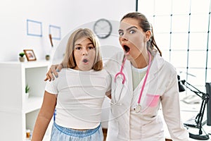 Pediatrician woman working at the clinic with little girl scared and amazed with open mouth for surprise, disbelief face