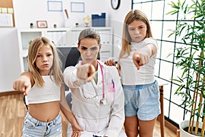 Pediatrician woman working at the clinic with two little girls pointing with finger to the camera and to you, confident gesture