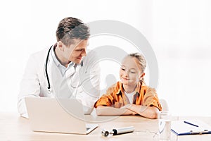 Pediatrist in white coat and child using laptop in clinic