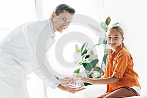 Pediatrist in white coat giving glass of water to child in clinic