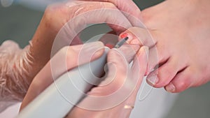 Pedicurist woman removes red gel shellac polish from toe using manicure machine.