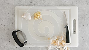 Peeled garlic, garlic press, and kitchen knife close-up on a white plastic cutting board, flat lay