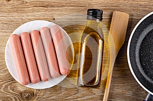 Peeled sausages in plate, bottle of vegetable oil, food turner, frying pan on table. Top view