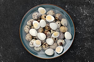 Peeled and unpeeled hard boiled quail eggs in plate on black table, top view