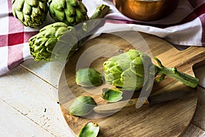 Peeling fresh ripe artichokes, preparing for cooking, round wood cutting board, knife, kitchen linen towel, copper dipper