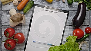 Pen rolling on blank clipboard on kitchen table with vegetables