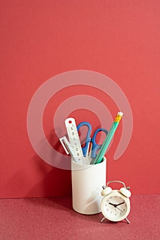 Pencil holder with ruler, scissors, pen and alarm clock on red desk. red wall background, workspace