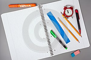 Pencils, a pen and a ruler lie on an open notebook. An alarm clock reminds of time.