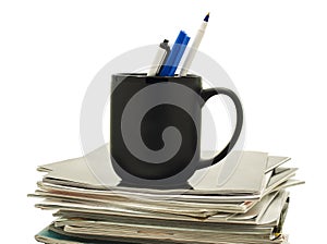 Pens in a coffee mug atop stack of magazines