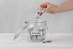 Pension concept. Woman putting dollar banknote into glass jar with money at white wooden table, closeup