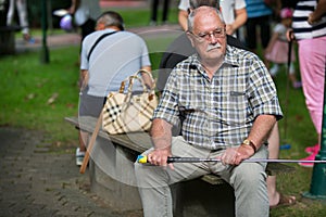 Pensioner at a minigolf court is waiting on a park bench and watching to their wife at a sunny day.