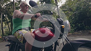 Pensioner riding motorcycle on tropical village at sunny day. Elderly man motorbiker driving on motorcycle on tropical