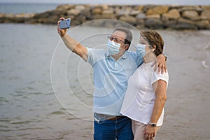 Pensioner woman and her husband taking romantic walk taking selfie - happy retired mature couple in face mask walking on the beach