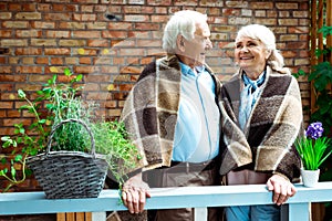 Pensioners smiling while standing in plaid blankets near flower pots