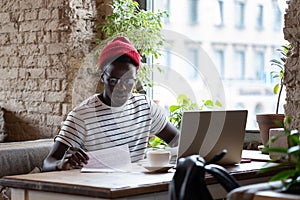 Pensive African American hipster man in glasses distance working on laptop with documents in cafe