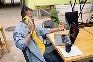 Pensive afro american businessman in stylish clothing talking on mobile phone and using laptop outdoors cafe, drinking